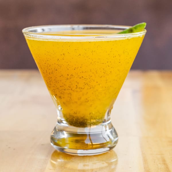 A vibrant yellow mango lassi cocktail in a martini glass, garnished with a green leaf.