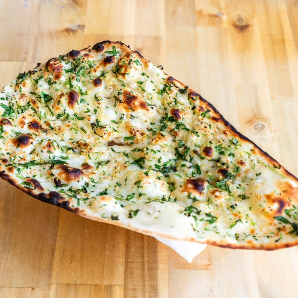 A close-up of a large, golden-brown garlic naan bread with fresh green herbs on a wooden surface.