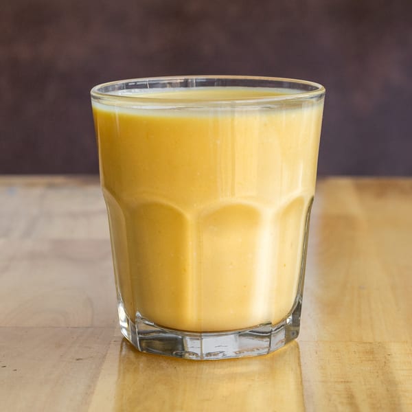 A glass of creamy, golden mango lassi, an Indian yogurt drink, on a light wooden table.