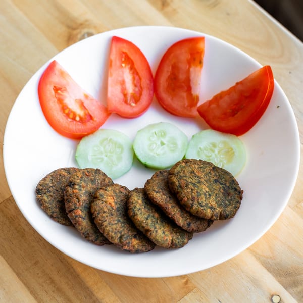 A white plate of Bombay Palace's Indian food with fried patties, tomato wedges, and cucumber slices.